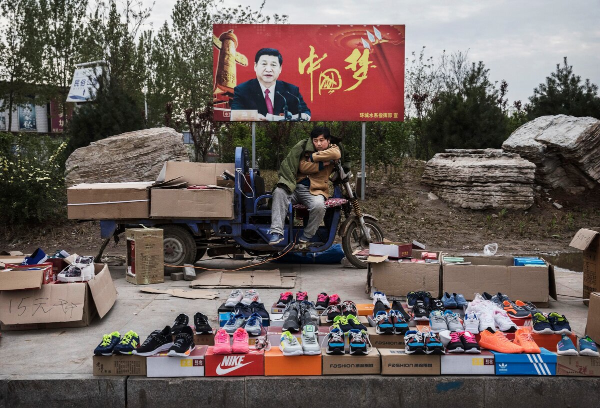 SHIJIAZHUANG, KÍNA - ÁPRILIS 9.: Egy kínai árus tornacipőket és cipőket árul az utcán egy Xi Jinping kínai elnököt ábrázoló, „China Dream” feliratú tábla előtt 2017. április 9-én a Hebei tartománybeli Shijiazhuangban, Kínában. Kína gazdasága 2017 első negyedévében 6,9 százalékkal nőtt - közölte a kormány 2017. április 17-i jelentése -, ami magasabb volt a vártnál, és újabb jel arra, hogy a világ második legnagyobb gazdasága stabilizálódhat (Fotó: Kevin Frayer/Getty Images).
