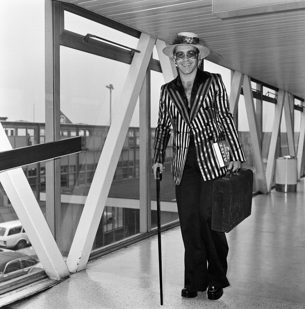 A popsztár Elton John virágokkal és tollakkal díszített kalapban érkezik a Heathrow repülőtérre Los Angelesből, ahol új albumát vette fel. Háromhetes tartózkodásra tért vissza, és prioritási listájának élén a holnapi Huddersfieldben megrendezésre kerülő labdarúgó-mérkőzés áll, ahol Elton kedvenc csapatának, a Watfordnak fog szurkolni, 1974. augusztus 30. (Fotó: Dennis Stone/Mirrorpix/Getty Images)
