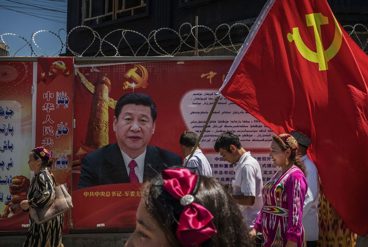 A Kínai Kommunista Párt etnikai ujgur tagjai zászlót visznek egy Xi-tábla mellett a kínai Kashgarban 2017 júniusában.